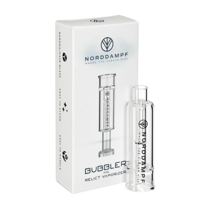 Auf dem Bild ist in Hintergrund die Originalverpackung des Bubbler/ Wasserfilter für den Norddampf Relict Kräuter Verdampfers zu sehen. Im Vordergrund steht der Wasserfilter aufrecht. Die Farbe des Glases ist transparent. Es handelt sich um ein Originales Zubehör für den Norddampf Relict Vaporizer.
